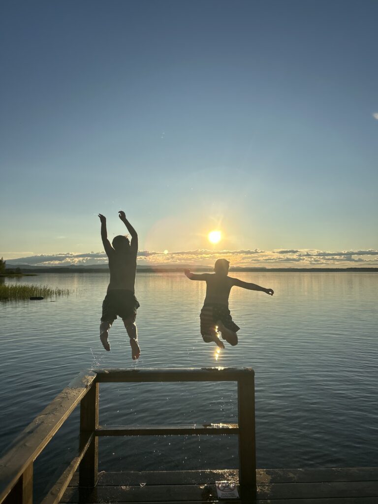 Två pojkar hoppar från en brygga, solnedgång. Foto.